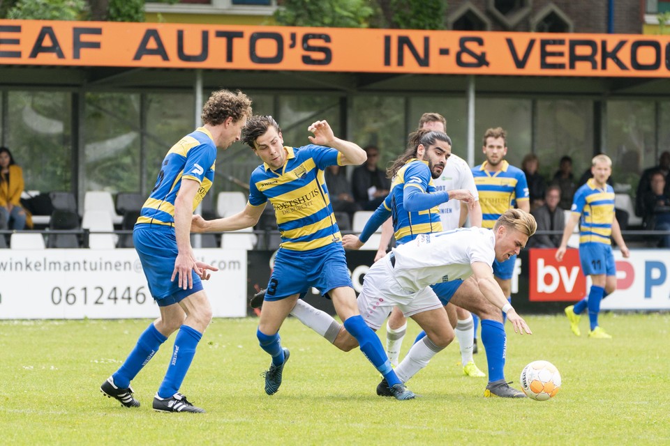 FC Boshuizen, in het geel en blauw, voetbalt tegen de achtergrond van een lange lage tribune.